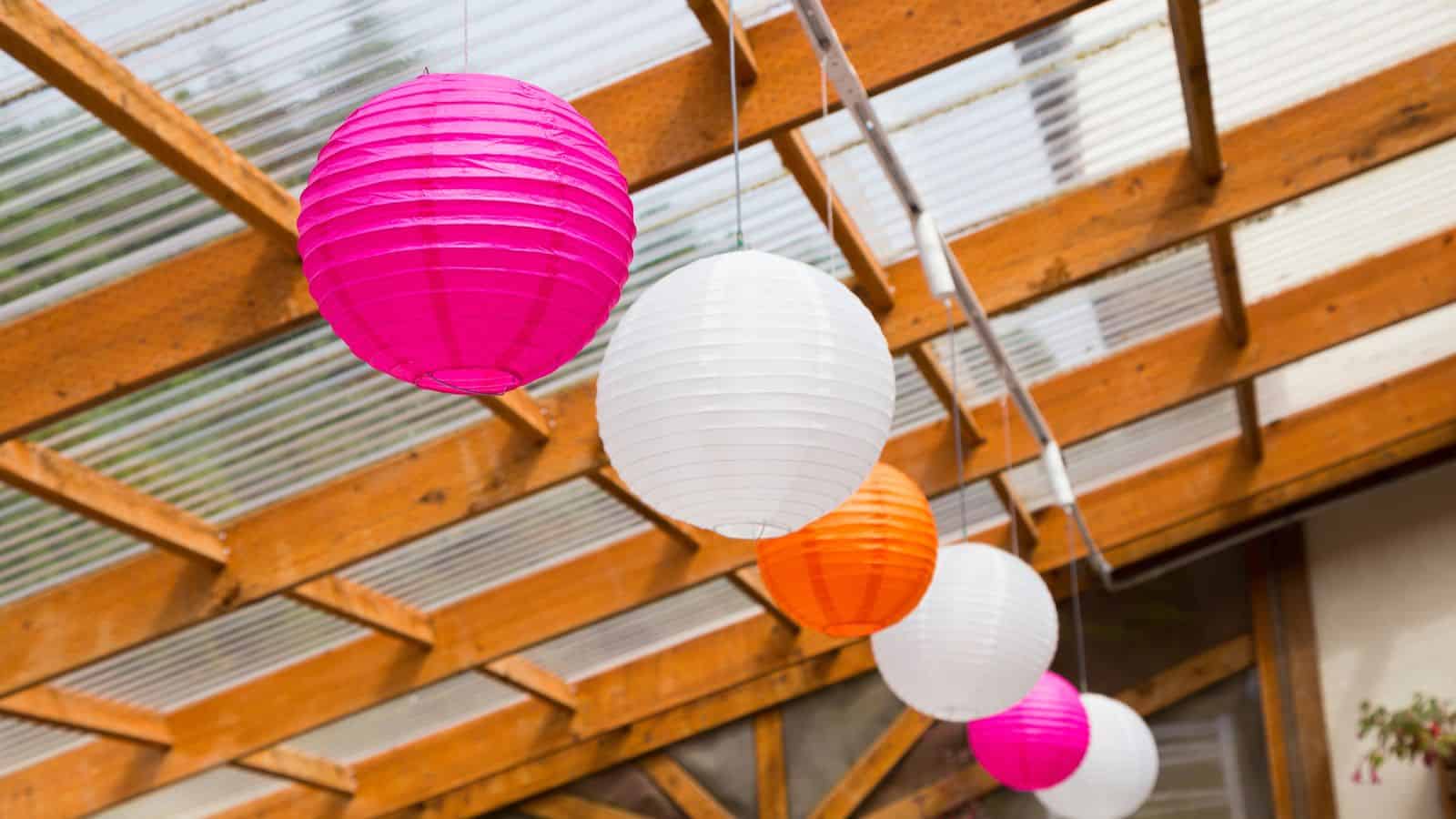Colorful paper lanterns, including pink, white, and orange, hang from a wooden pergola with a transparent roof. The background shows a partial view of a building.