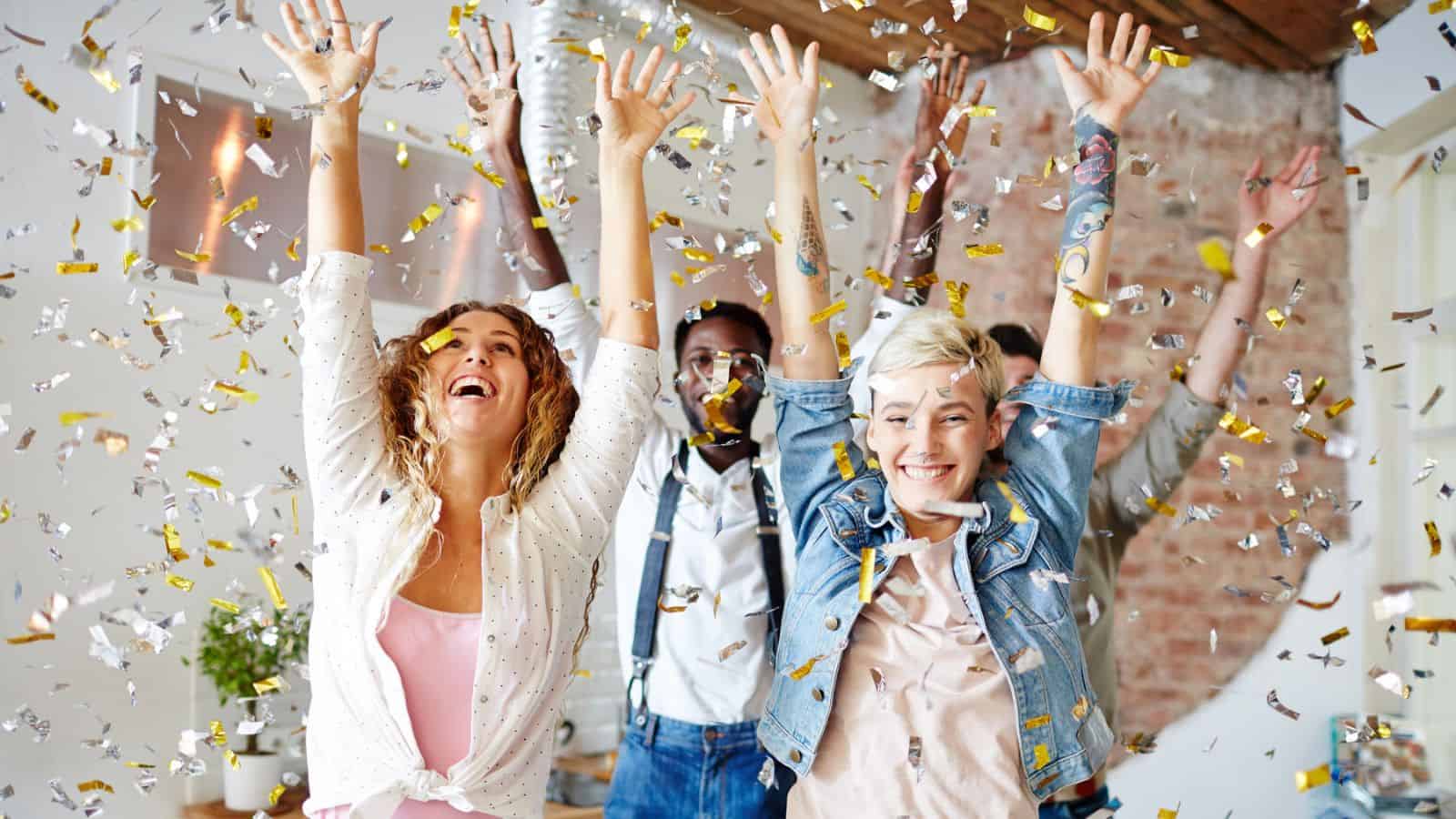 A group of five people indoors celebrating with arms raised, surrounded by falling confetti. They seem joyful and excited, standing in a space with exposed brick walls and plants in the background.
