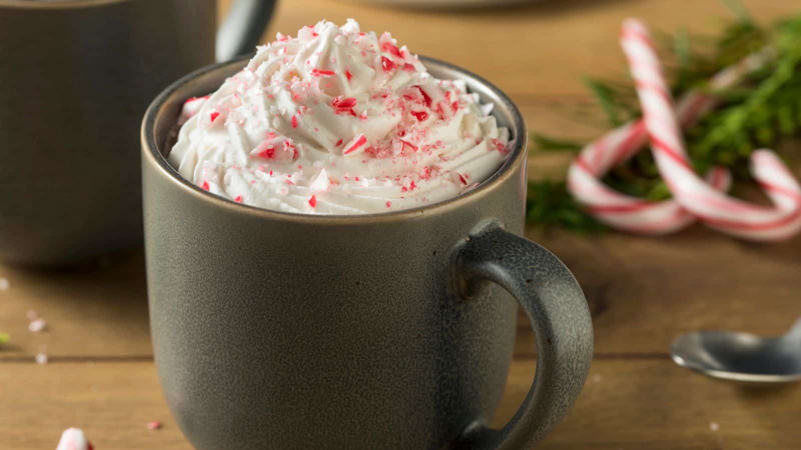 A cup of hot chocolate topped with whipped cream and crushed candy canes sits on a wooden table. In the background, blurred candy canes and greenery are visible, along with part of another cup and a spoon.