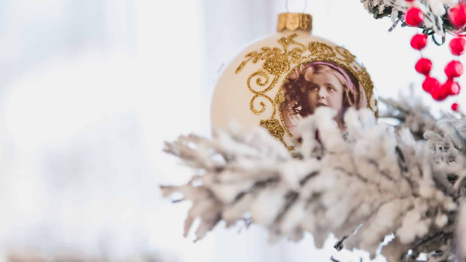 A Christmas tree ornament hangs from a snowy branch. The ornament is round, with a decorative golden pattern and an image of a person on its surface. Red berries are visible in the background, adding to the festive scene.