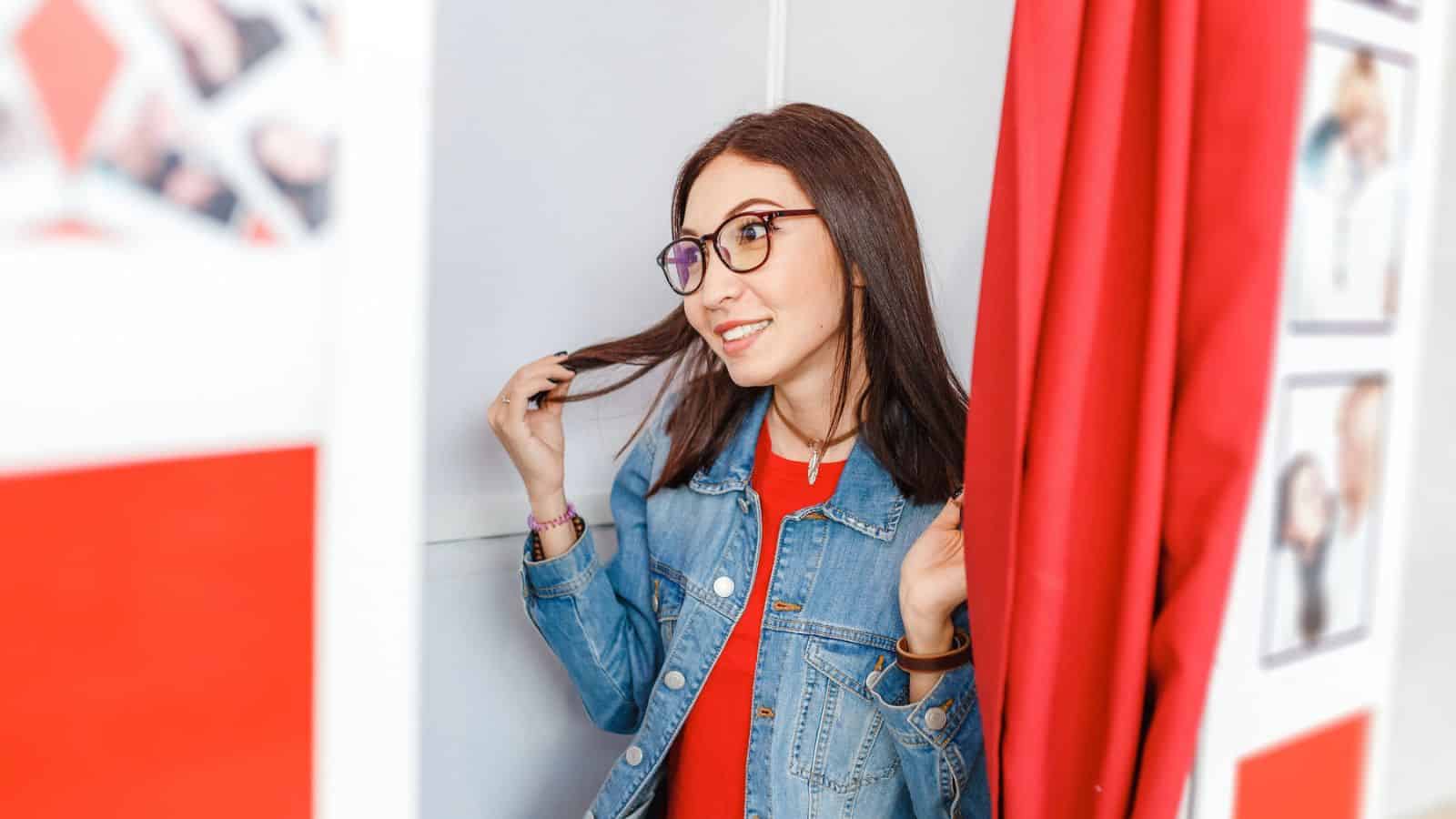 A person with long dark hair and glasses, wearing a denim jacket and red shirt, stands partially behind a red curtain. They are smiling and holding a strand of hair. The background includes blurred images on the wall.
