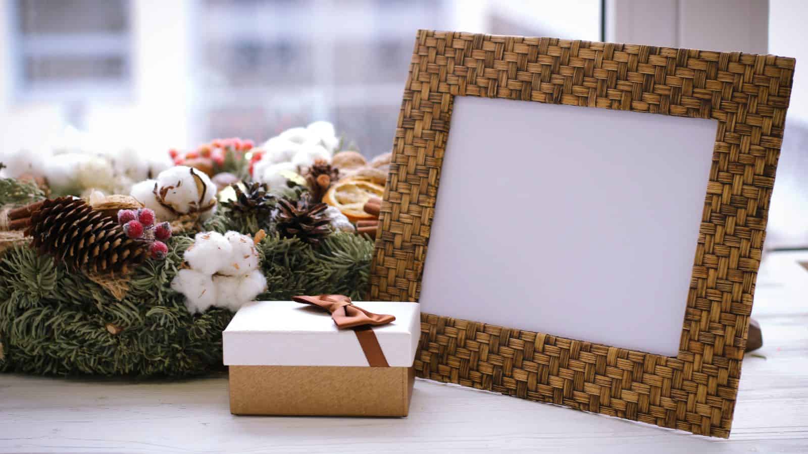 A woven wooden picture frame is displayed next to a small gift box with a brown ribbon. In the background, there is a decorative wreath made of cotton, pinecones, and greenery on a windowsill.