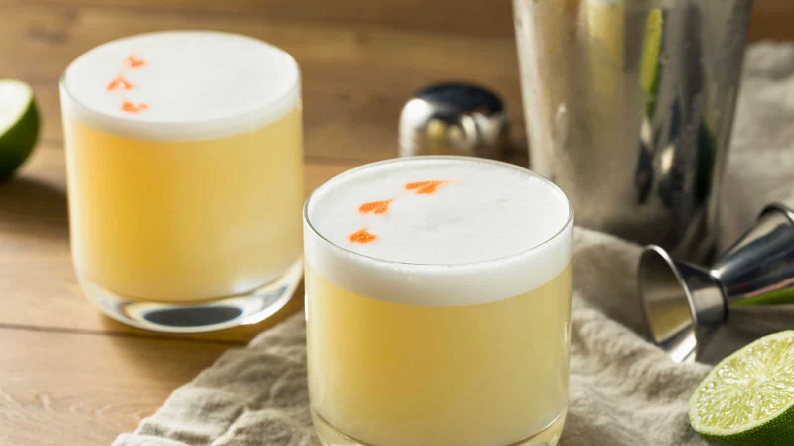 Two frothy cocktails in glass tumblers sit on a cloth with lime wedges nearby. The drinks have a white foam top with a few drops of orange bitters. A cocktail shaker and jigger are in the background.