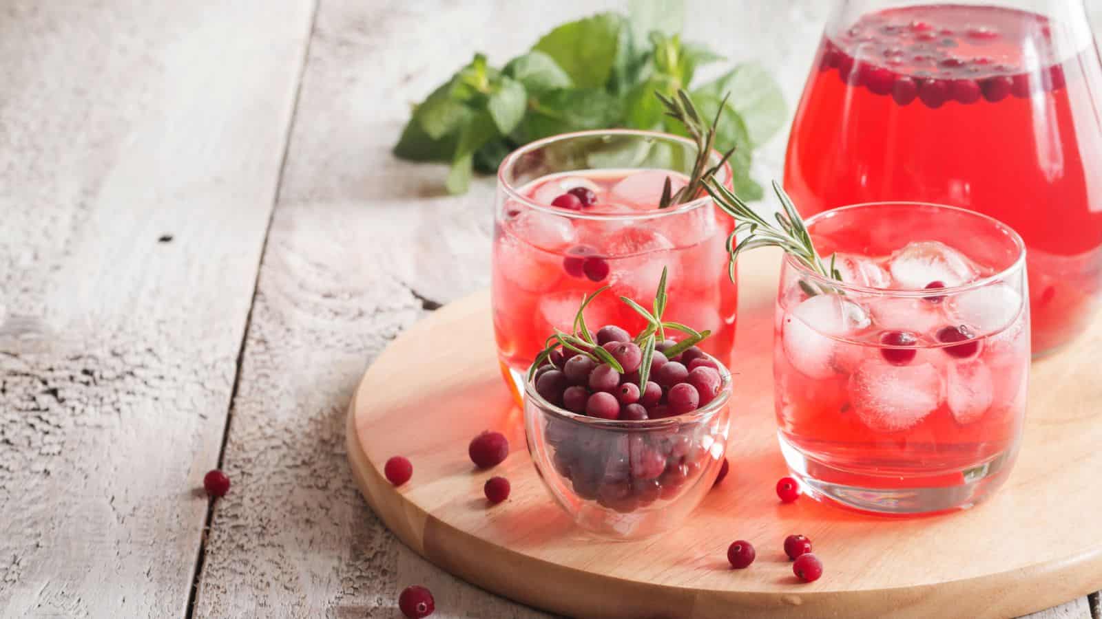 Three glasses of cranberry juice with ice and rosemary garnish sit on a wooden tray next to a small bowl of cranberries and a pitcher filled with juice. A sprig of mint is placed in the background on a rustic wooden surface.