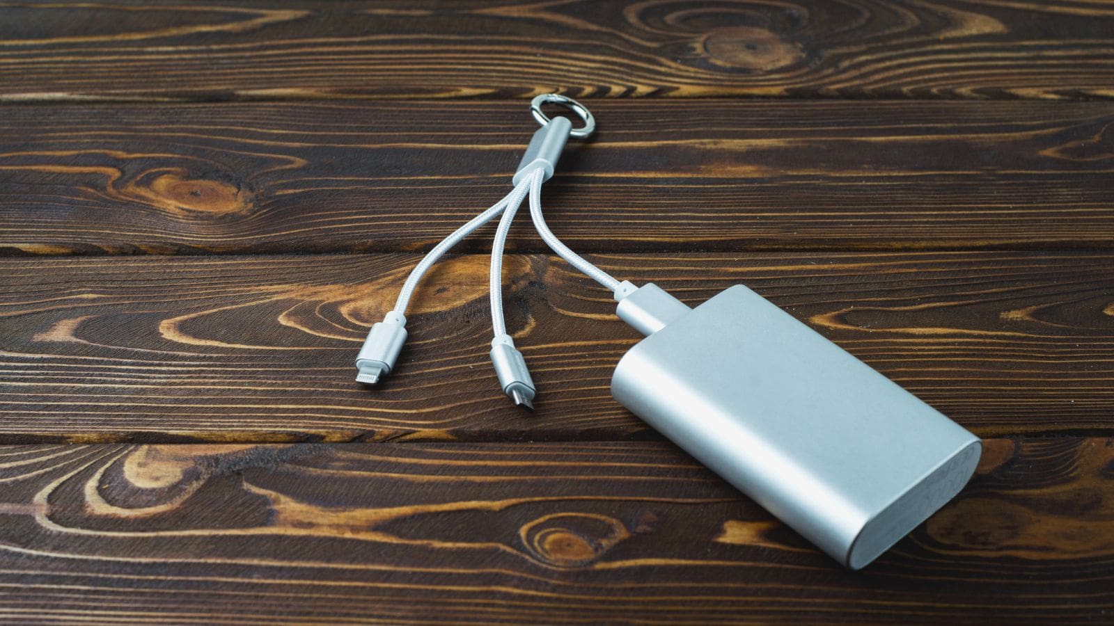 A silver power bank with a keyring and three attached charging cables is placed on a wooden surface. The cables have different connectors: USB-C, Lightning, and Micro USB.