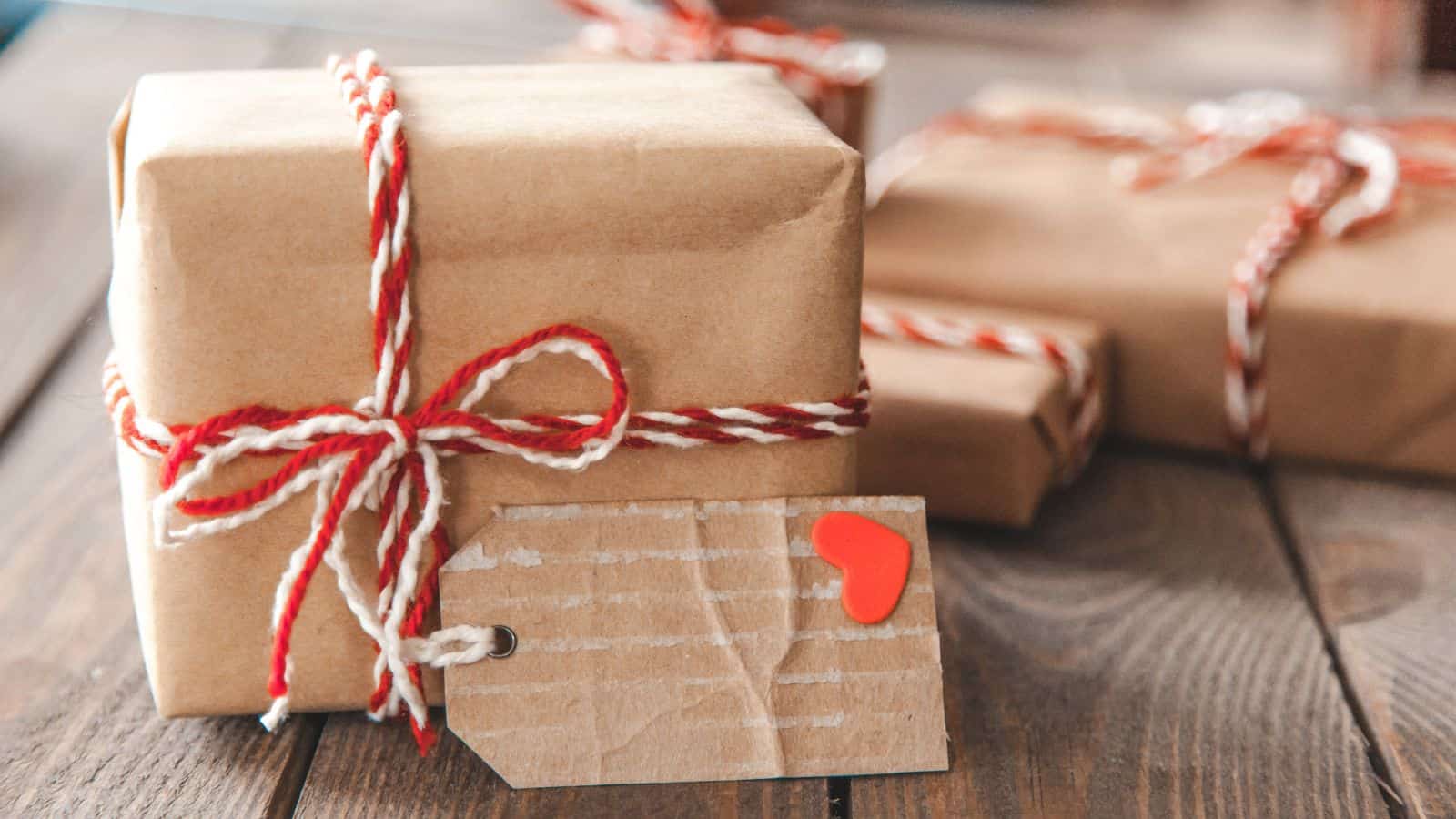 A small gift wrapped in brown paper with a red and white twine bow sits on a wooden surface. Attached is a brown tag with a red heart sticker. In the background, two more similarly wrapped gifts are slightly blurred.