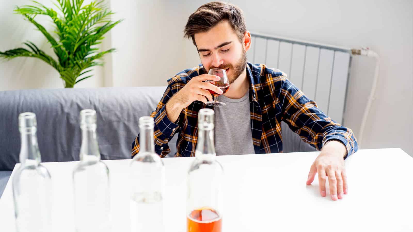 A man sits at a table, holding a glass of brown liquid, seemingly lost in thought. Several empty bottles are lined up in front of him. A green plant and a radiator are visible in the background.