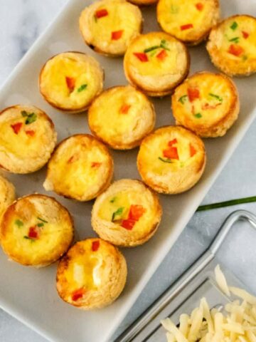 A rectangular white plate with multiple mini quiches containing visible diced bell peppers and herbs. Nearby, some sliced bread and a small pile of grated cheese are on a silver tray. Green chives are scattered on a marble surface.
