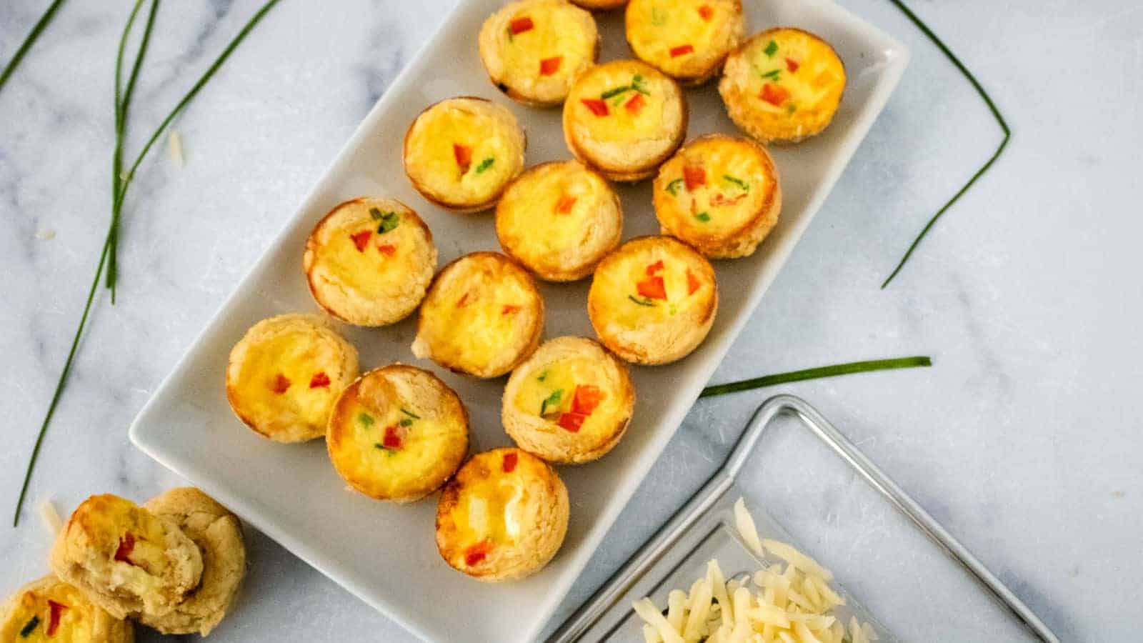 A rectangular white plate with multiple mini quiches containing visible diced bell peppers and herbs. Nearby, some sliced bread and a small pile of grated cheese are on a silver tray. Green chives are scattered on a marble surface.