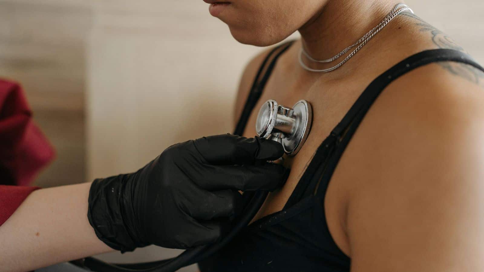 A healthcare professional wearing black gloves uses a stethoscope to listen to a person's chest. The person is wearing a black tank top and a necklace.
