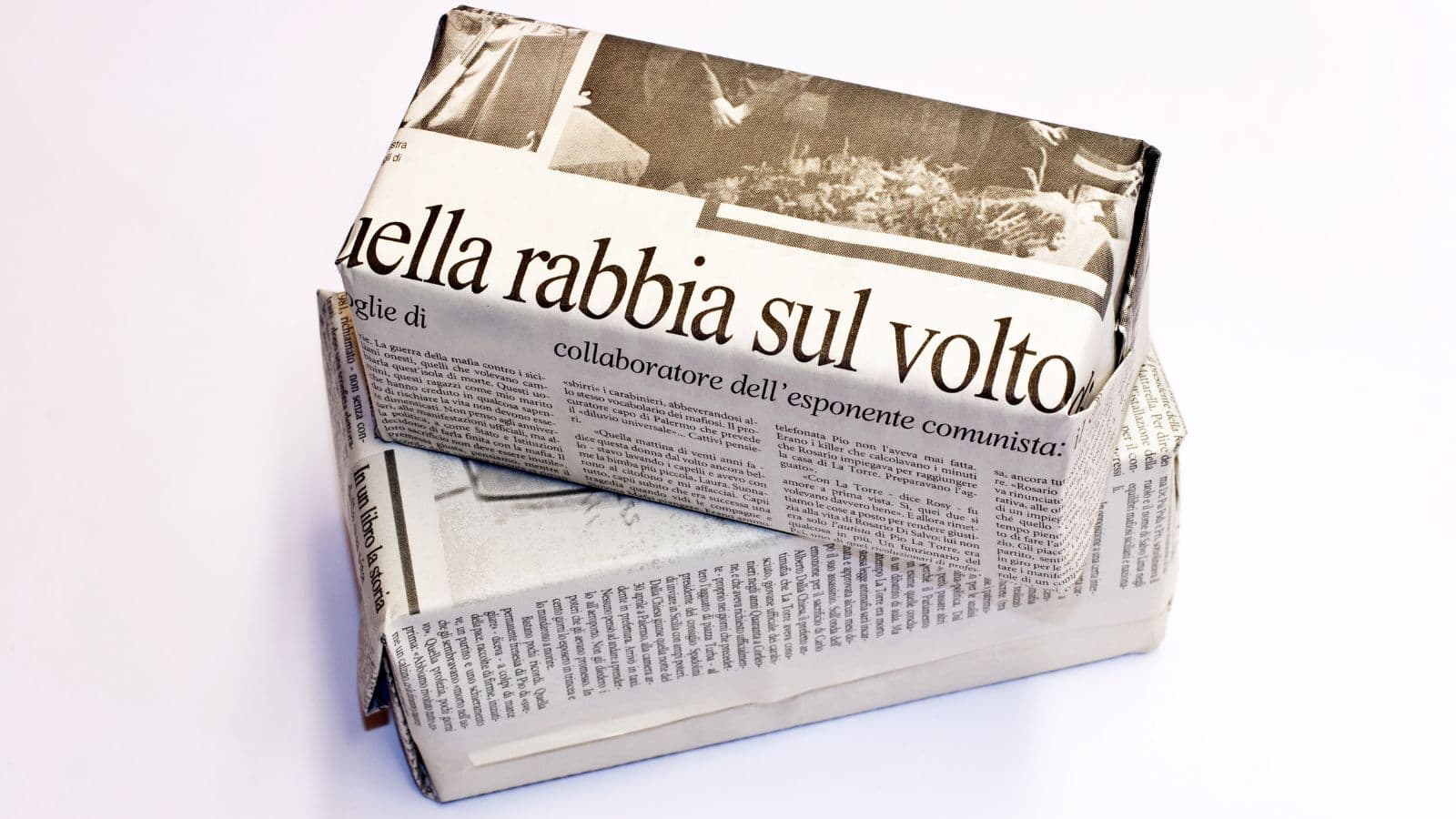 Two rectangular packages wrapped in newspaper are stacked on top of each other against a plain white background. The newspaper text is in Italian and includes a visible headline.