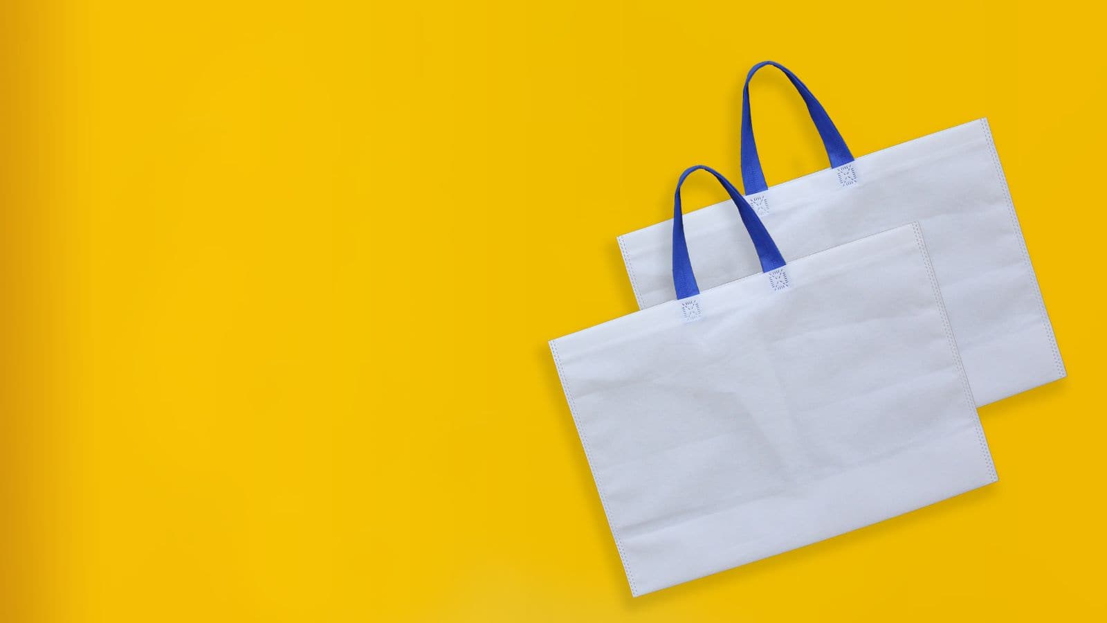Two white tote bags with blue handles are placed against a bright yellow background. The bags are positioned at an angle, overlapping slightly, with the handles facing upwards.