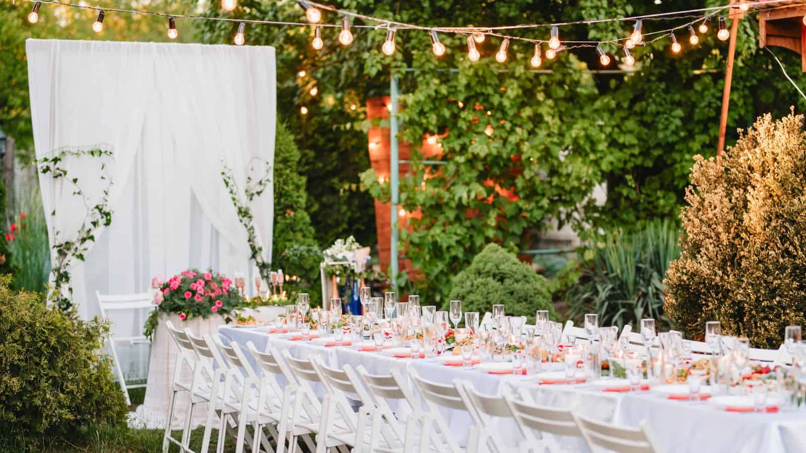 A long, decorated table set for an outdoor event, surrounded by white chairs. The table is adorned with tableware and flower arrangements. String lights are hung above, and greenery surrounds the area, creating a festive atmosphere.