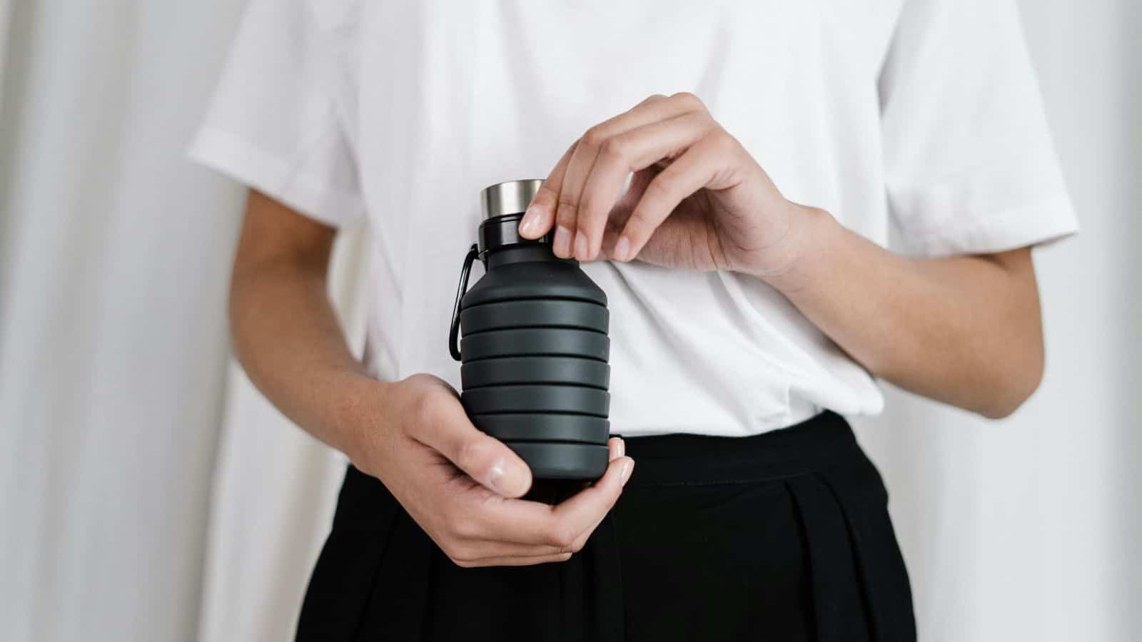 A person wearing a white t-shirt and black pants is holding a small, black, ribbed water bottle with a metal cap. The focus is on the hands and the bottle against a plain background.