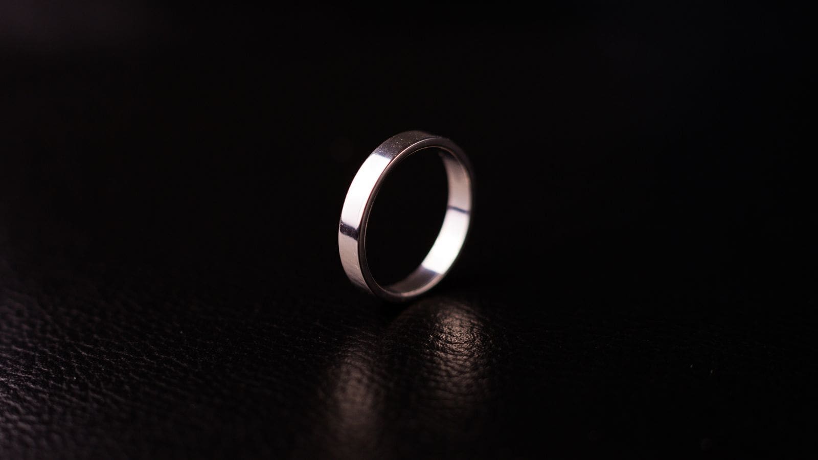 A silver ring is standing upright on a black, textured surface with light reflecting off its smooth surface. The dark background contrasts with the shiny metal of the ring.