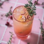 A pink cocktail garnished with cranberry slices, a lemon slice, and a sprig of thyme is served in a ribbed glass on a pink tiled surface. Additional thyme sprigs and a few cranberries are scattered around the glass.