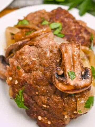 A plate with a cooked Salisbury steak topped with sliced mushrooms and garnished with fresh parsley. The meal is placed on a wooden surface with some greens in the background.