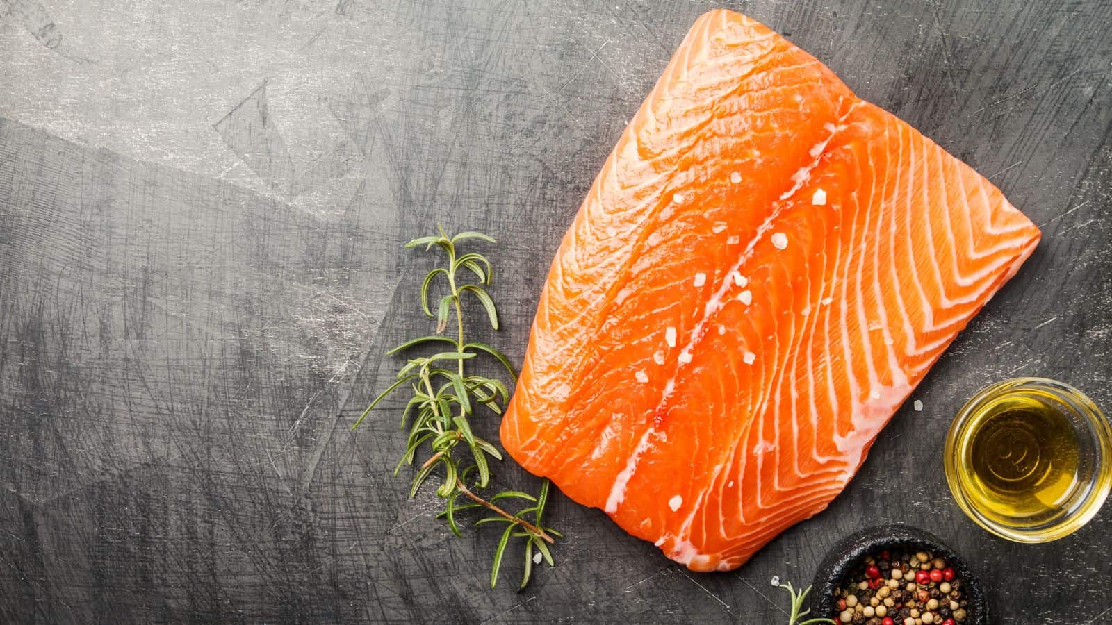 A raw salmon fillet is placed on a dark surface next to a small bowl of mixed peppercorns, a sprig of rosemary, and a bowl of oil. The fish is bright orange with visible striations, and salt crystals are sprinkled on top.