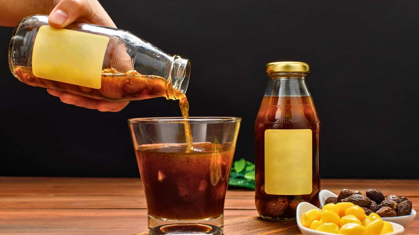 A hand pours a brown liquid from a bottle with a blank yellow label into a glass. Another similar bottle and a bowl of yellow and dark round objects are on a wooden surface against a black background.
