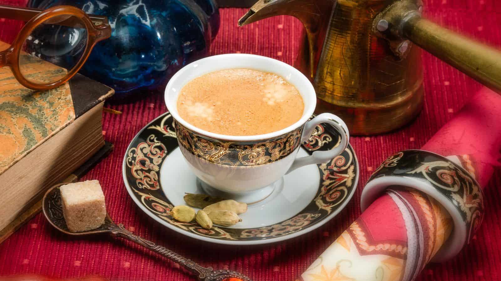 A decorated cup of coffee sits on a matching saucer, surrounded by cardamom pods. Nearby are a spoon with a sugar cube, a rolled scroll, a stack of books, an oil can, blue glassware, and eyeglasses, all on a red textured surface.