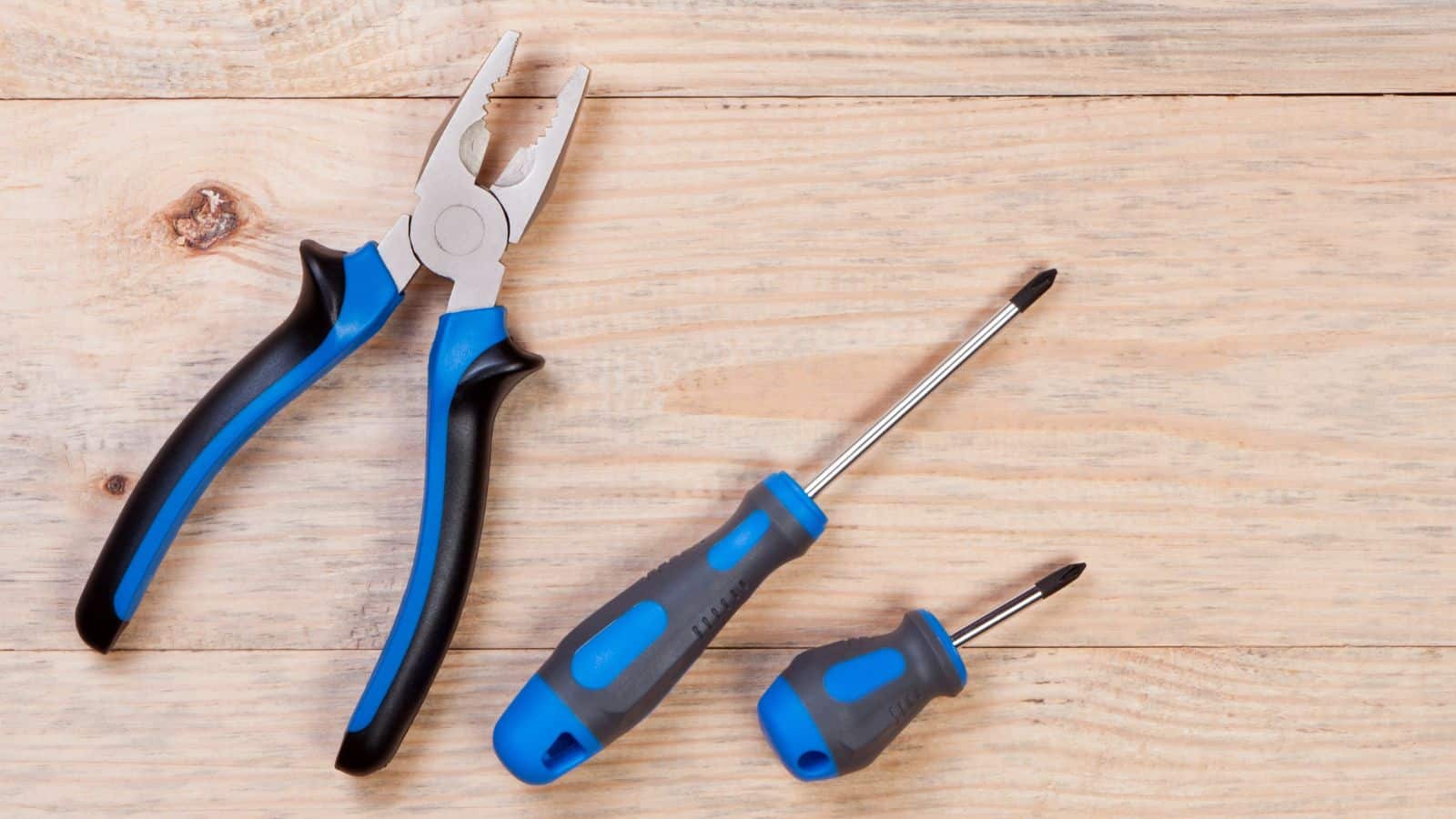 A pair of pliers and two screwdrivers with blue and black handles are placed on a wooden surface. The screwdrivers include one Phillips head and one flathead.