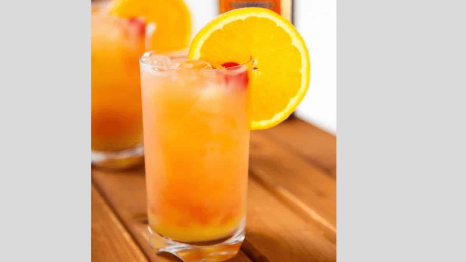 A tall glass filled with an orange cocktail, garnished with a slice of orange on the rim, is placed on a wooden surface. Another similar drink is partially visible in the background.