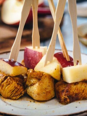 A plate with skewers featuring pieces of cooked chicken and apple chunks. The skewers are arranged upright on the plate. In the background, there are blurred kitchen items and ingredients.