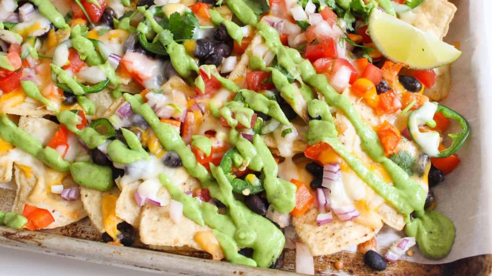 A tray of loaded nachos topped with melted cheese, black beans, diced tomatoes, red onions, jalape&ntilde;os, and drizzled with green sauce. A lime wedge and cilantro garnish the dish.