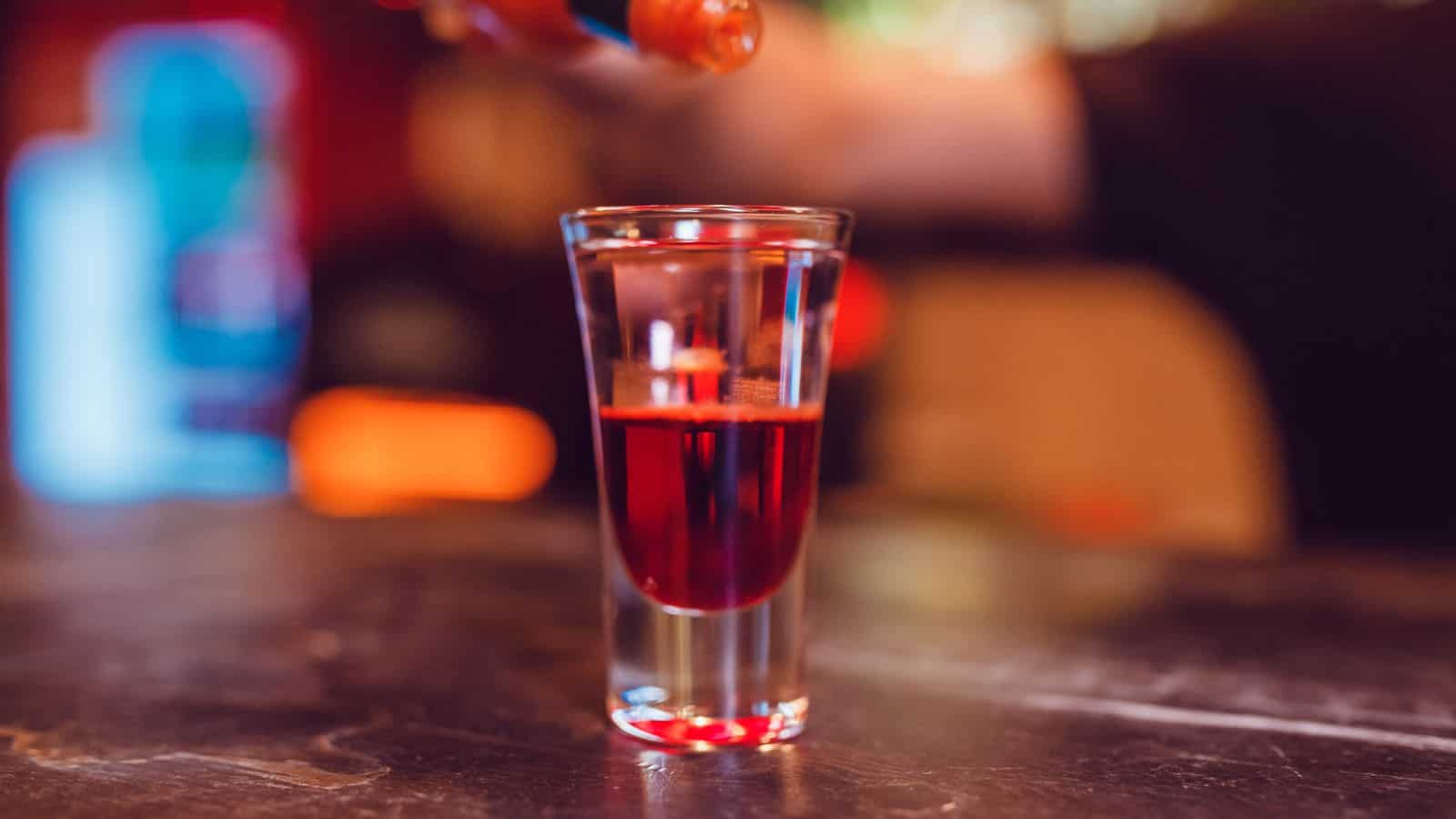 A close-up of a shot glass filled with a red liquid, placed on a bar counter. A hand is visible in the background, holding a small bottle above the glass. The environment appears to be a dimly lit bar or pub.