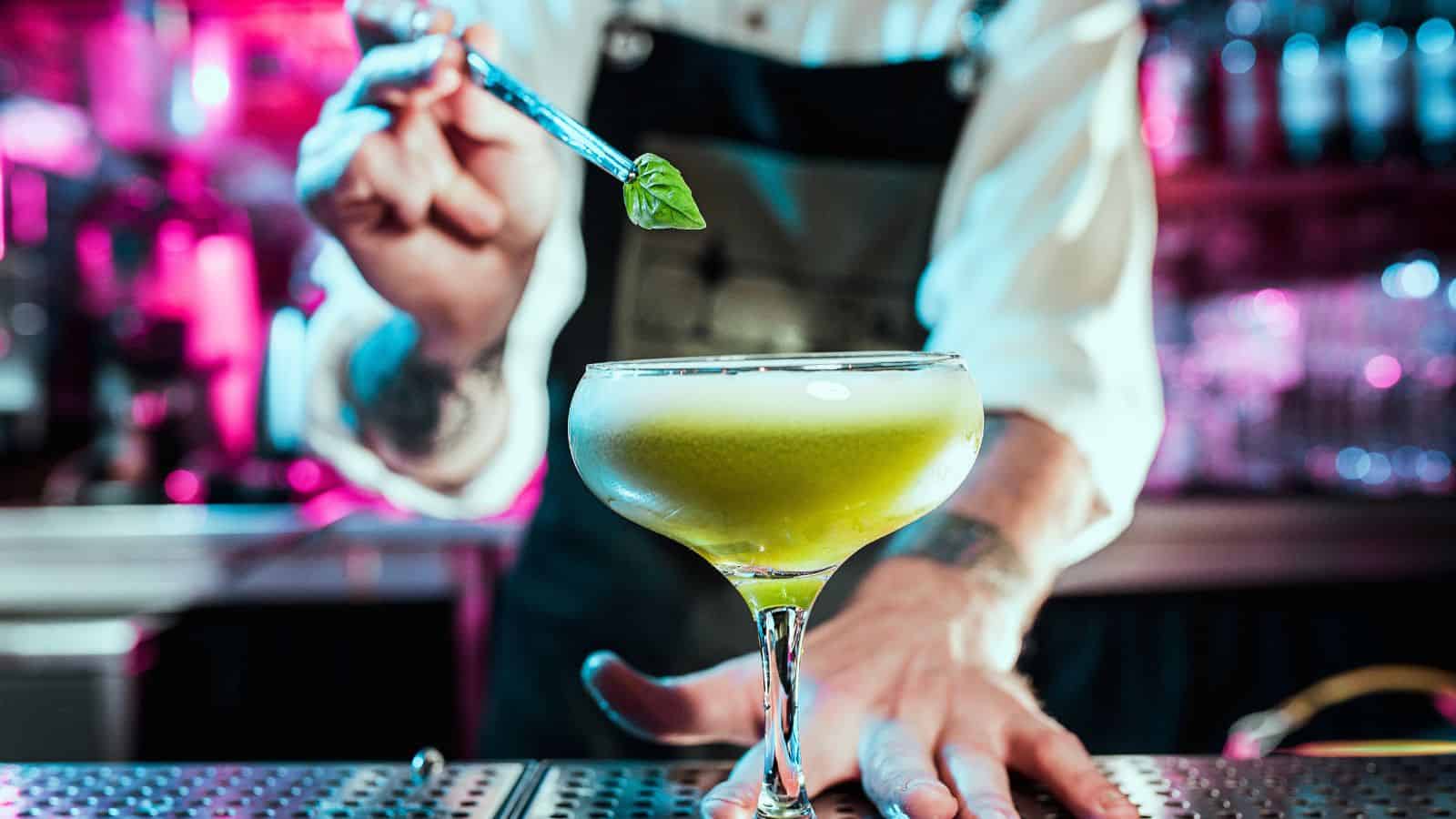 A bartender garnishes a light green cocktail in a coupe glass with a small leaf. The scene is set in a bar with blurred pink and purple lighting in the background. The bartender's arm tattoo is partially visible.