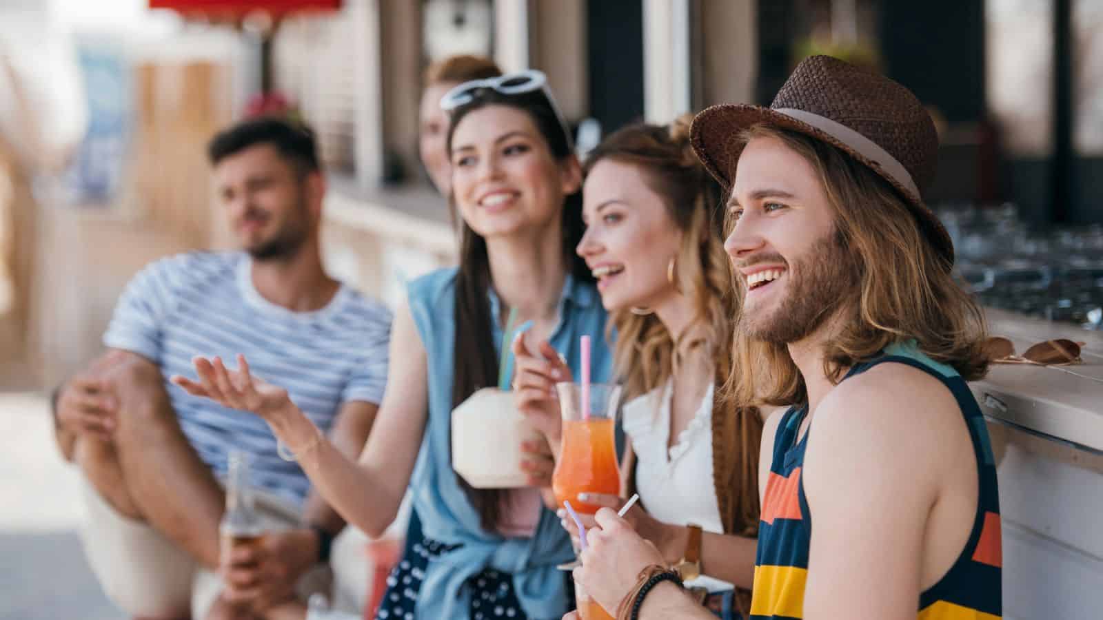 A group of four people sitting outdoors, enjoying drinks. One person wears a hat and striped shirt, holding a cocktail. Another wears sunglasses on their head and holds a coconut drink. They appear to be laughing and having a good time.