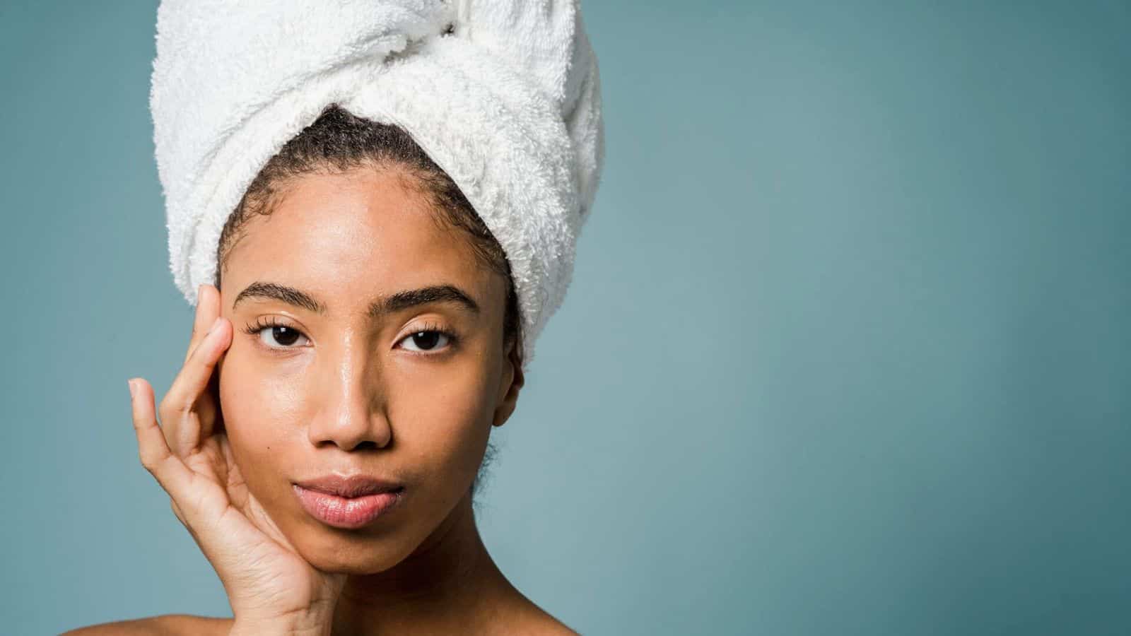 A person with a white towel wrapped around their head is touching their face with one hand. Their expression is calm and the background is a simple blue.