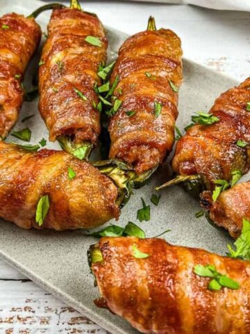A rectangular plate holds seven bacon-wrapped jalapeños, garnished with chopped parsley. The jalapeños are evenly spaced on a wooden surface, and the bacon is cooked to a golden brown.