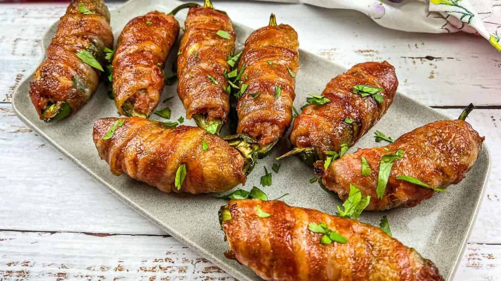 A rectangular plate holds seven bacon-wrapped jalape&ntilde;os, garnished with chopped parsley. The jalape&ntilde;os are evenly spaced on a wooden surface, and the bacon is cooked to a golden brown.