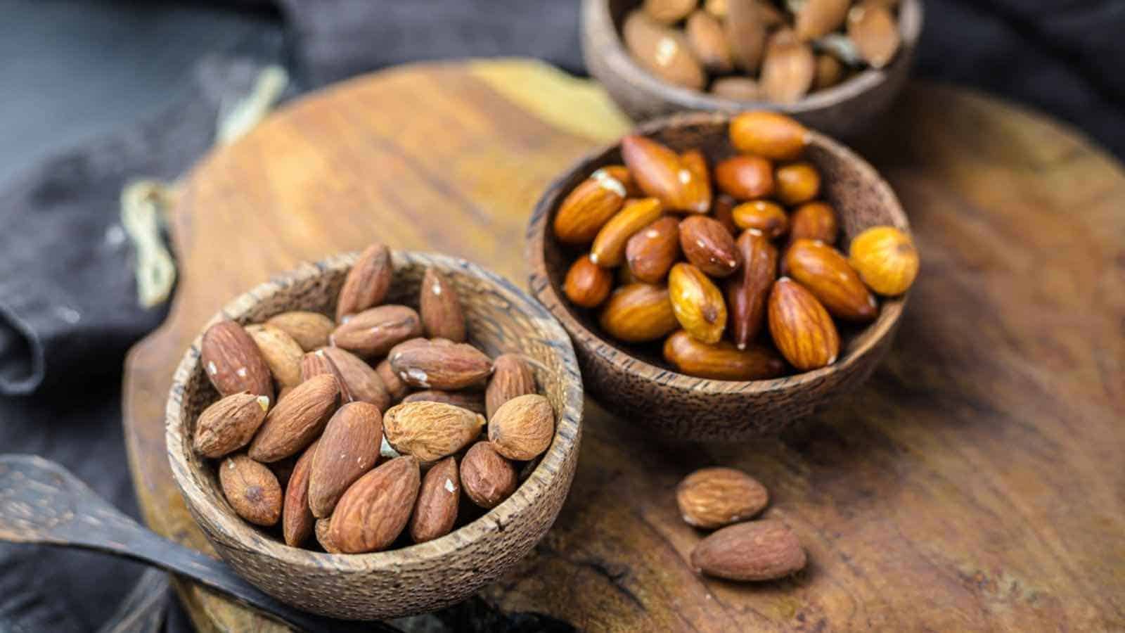 Three wooden bowls filled with different types of nuts are placed on a wooden surface. The nuts include almonds and possibly other varieties. The background is slightly blurred, featuring a dark cloth.