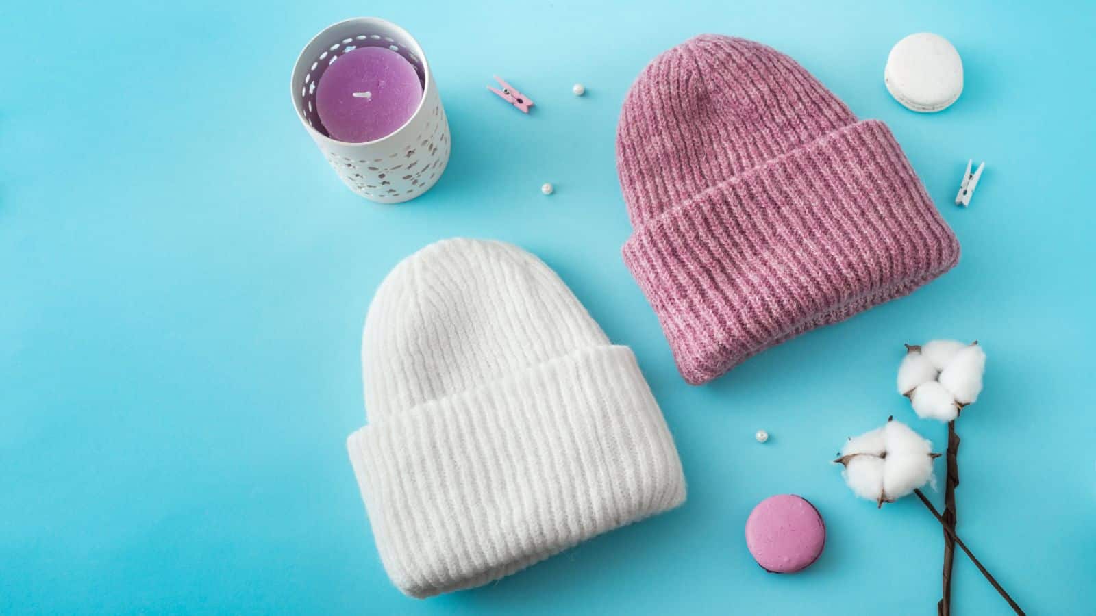 Two knit beanies, one white and one pink, are placed on a blue surface. Nearby are a purple candle in a holder, cotton sprigs, small white and pink macarons, and two tiny clothespins. Small white beads are scattered around.