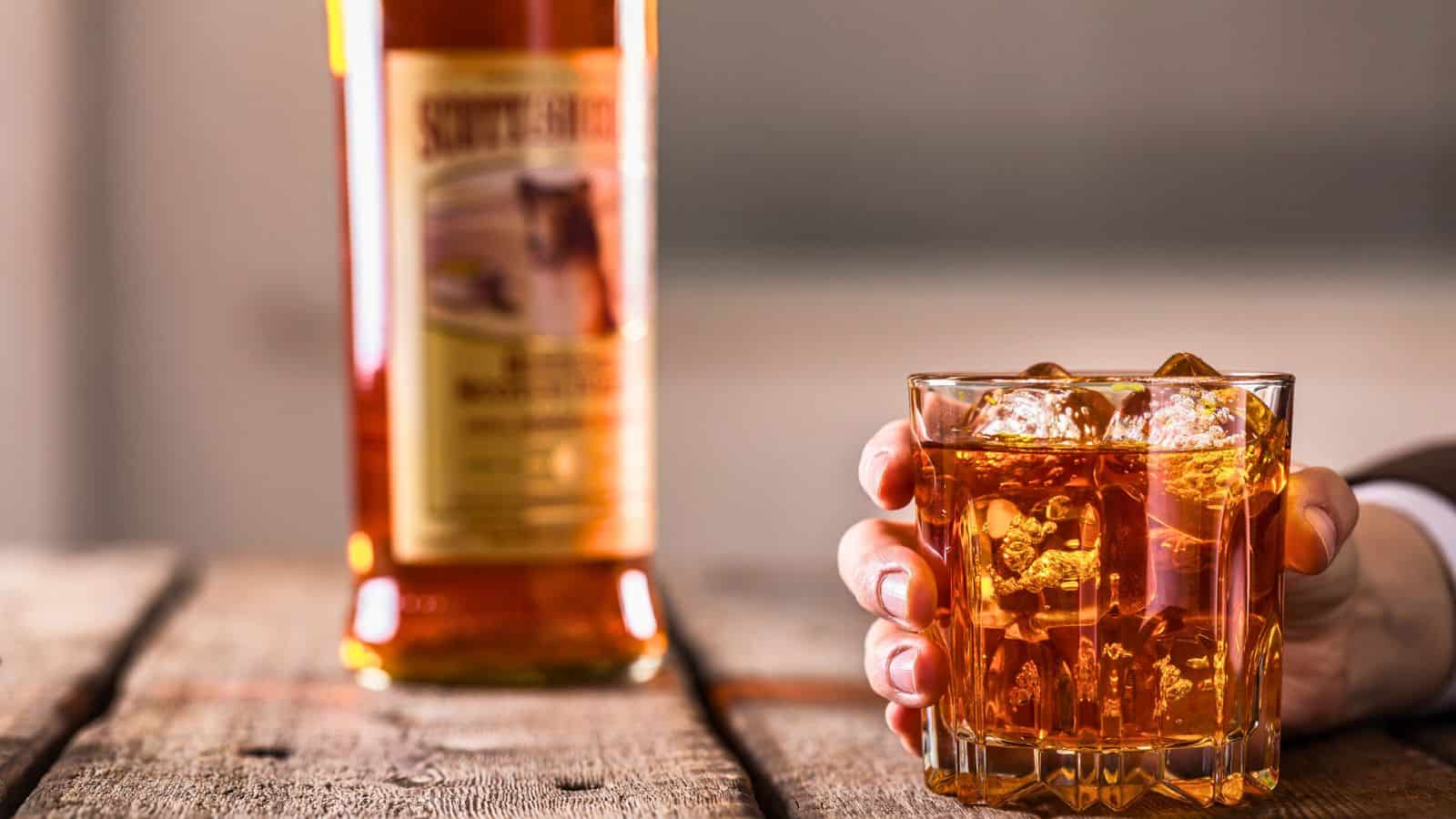 A hand holding a glass of whiskey with ice cubes is placed in the foreground on a wooden table. The background shows a bottle of whiskey slightly out of focus.