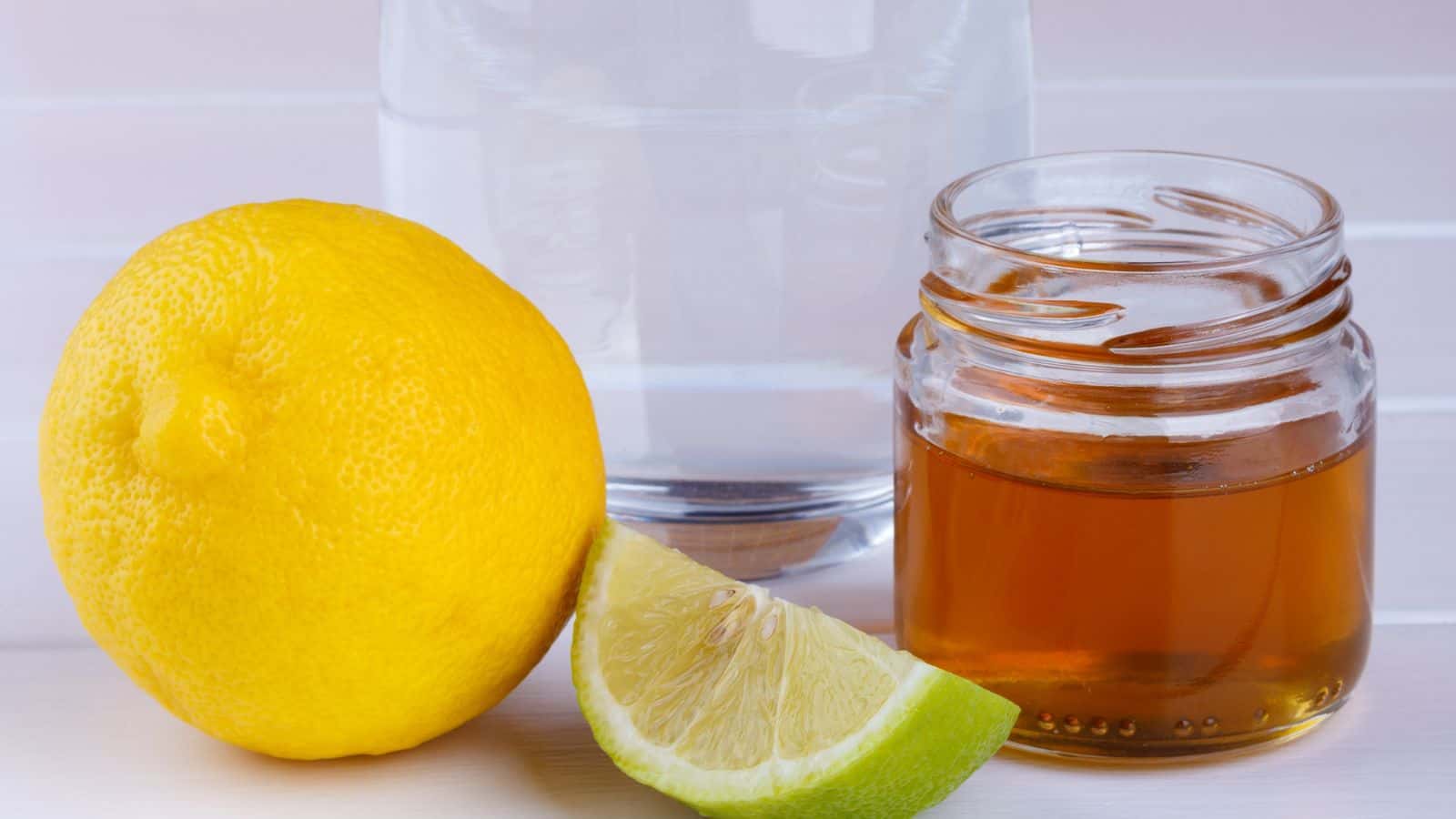 A whole lemon and a lemon wedge are placed beside a small glass jar of honey. Behind them is a glass of water. The objects are arranged on a light-colored surface.