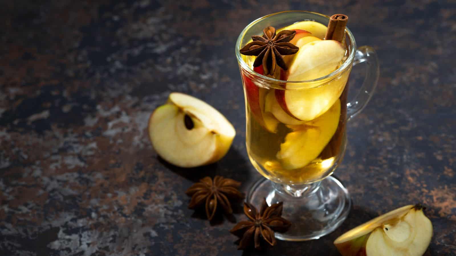 A glass mug filled with a hot beverage, garnished with apple slices, star anise, and a cinnamon stick, is placed on a textured dark surface. Two apple wedges and additional star anise are scattered around the mug.
