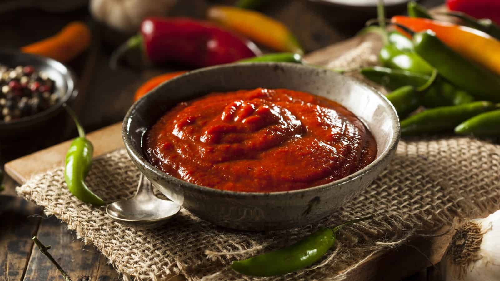 A bowl of red chili sauce sits on a burlap cloth, surrounded by green and red chili peppers. A metal spoon rests in the bowl, and the setting is a rustic wooden table.