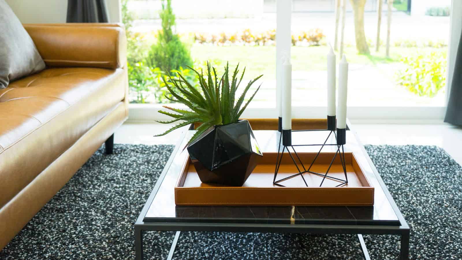 A modern living room scene featuring a brown leather sofa partially visible on the left, a black geometric vase with a green plant on a tray, and a candle holder with two white candles on a rectangular coffee table with a dark marble surface.