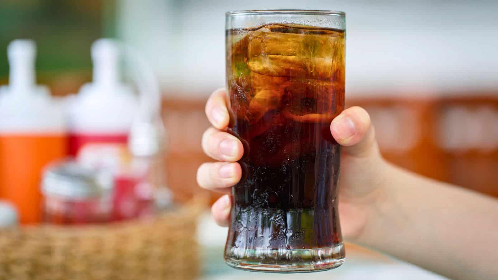 A hand holding a tall glass filled with dark, carbonated soda and ice cubes. In the background, there are blurred condiment containers on a table.