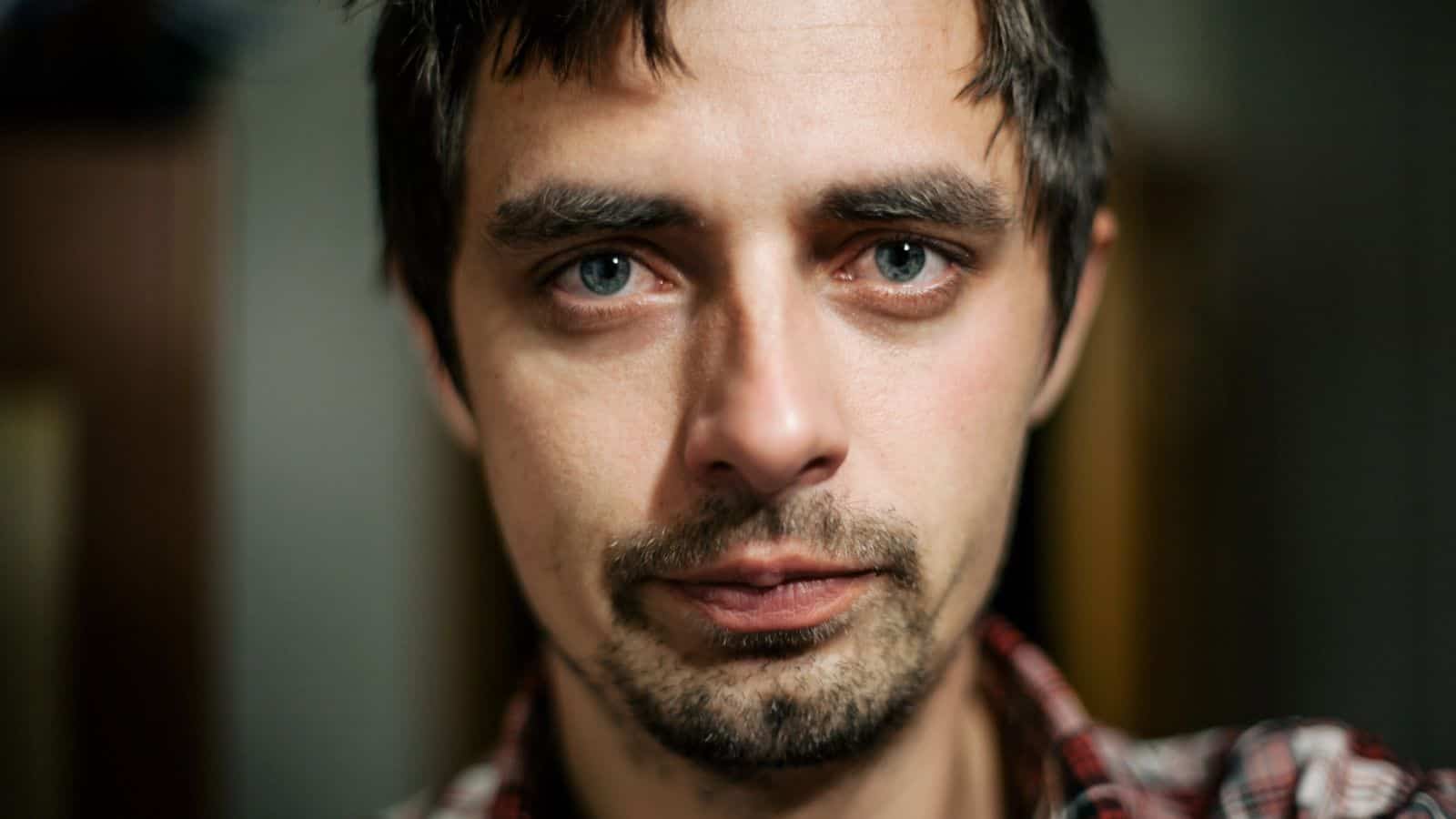 A man with short dark hair and a beard is looking directly at the camera. He is wearing a red plaid shirt. The background is blurry, emphasizing his facial expression.