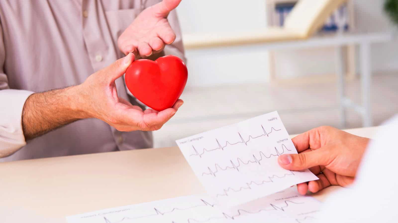 A person holds a red heart-shaped object while another person holds ECG prints. The scene is set in a medical environment, suggesting a discussion about heart health.
