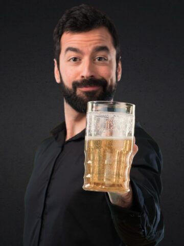 A man with a beard is holding a large glass of beer in an outstretched hand. He is wearing a black shirt and standing against a dark background.