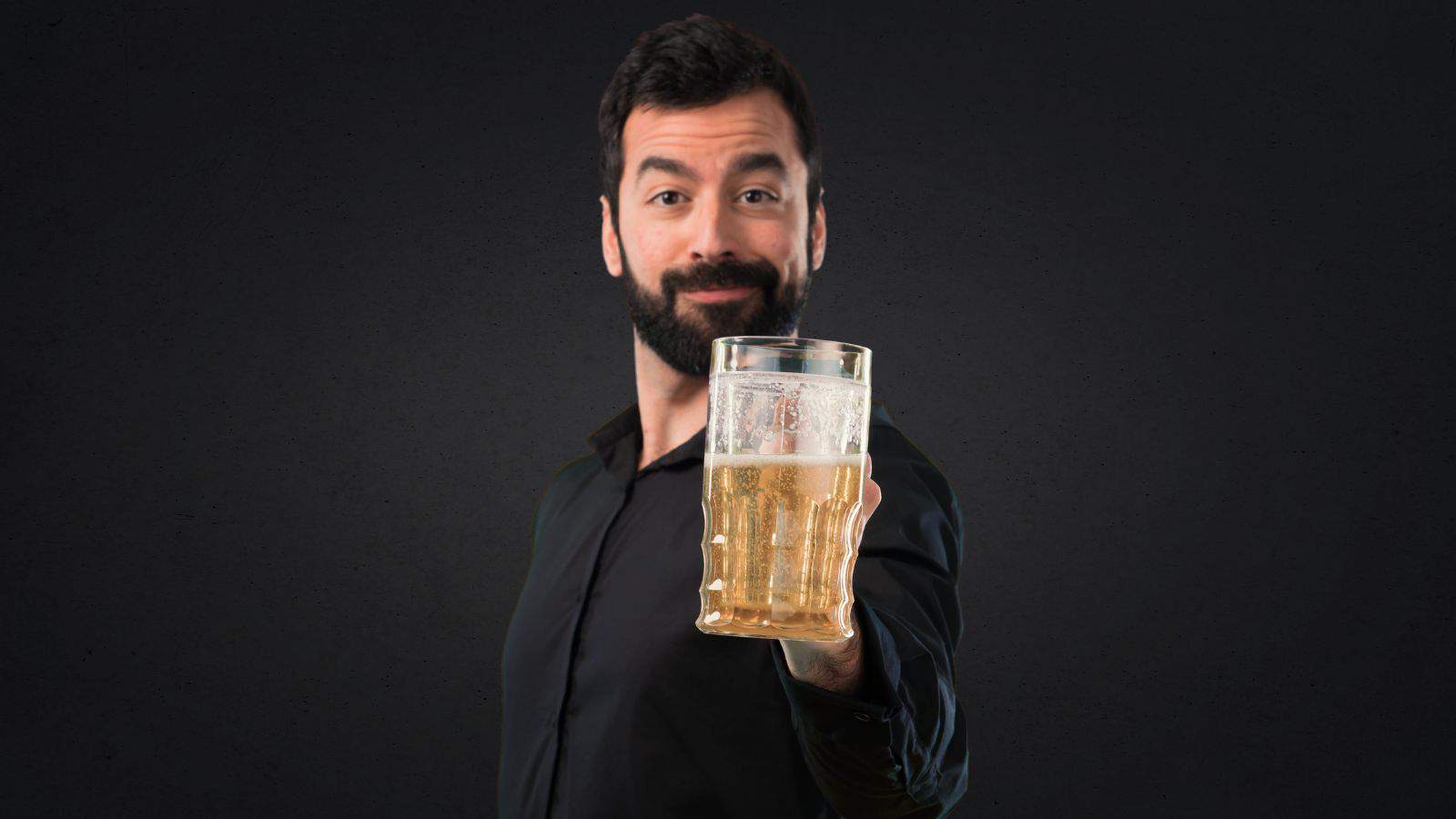 A man with a beard is holding a large glass of beer in an outstretched hand. He is wearing a black shirt and standing against a dark background.