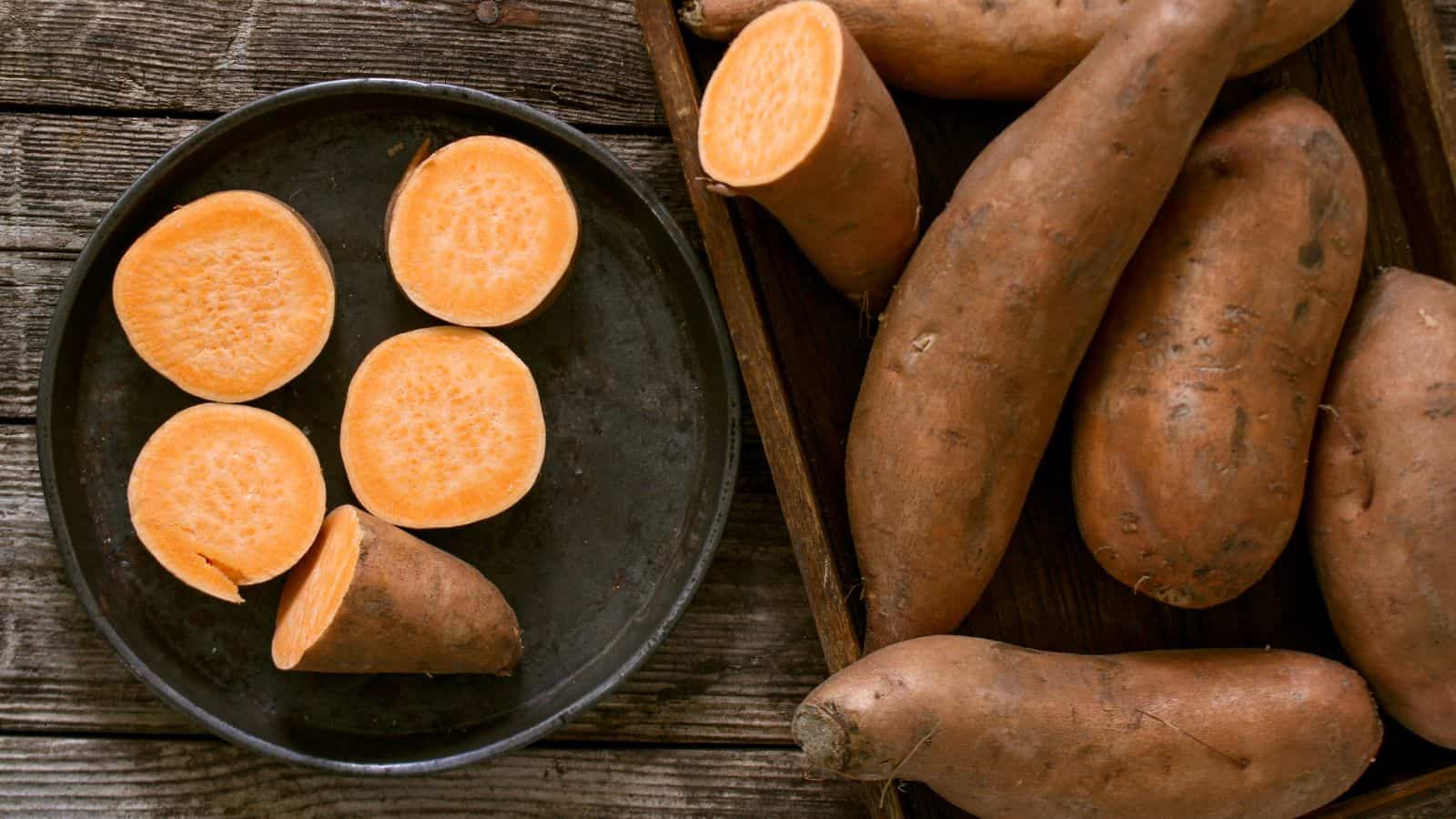 A black plate with five orange sweet potato slices is placed on a wooden surface. Next to the plate, several whole unpeeled sweet potatoes are arranged in a wooden tray. The background consists of rustic wooden planks.