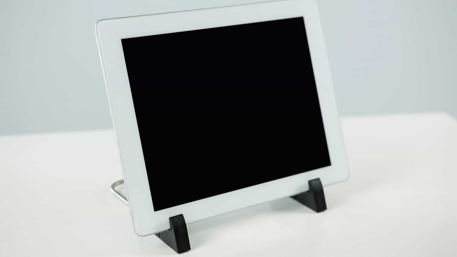 A white tablet with a blank black screen sits upright on a white surface, supported by a black stand. The background is a soft grayish color.