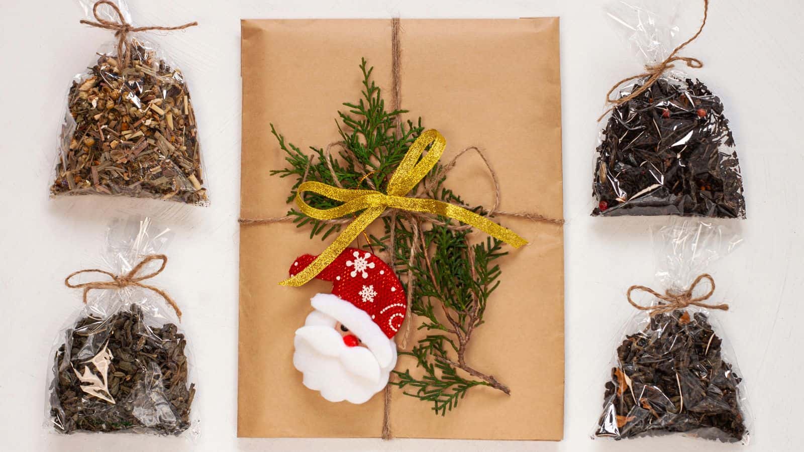 A wrapped gift decorated with green foliage, a gold ribbon, and a Santa Claus ornament is surrounded by five small bags filled with dried herbs tied with twine on a white background.