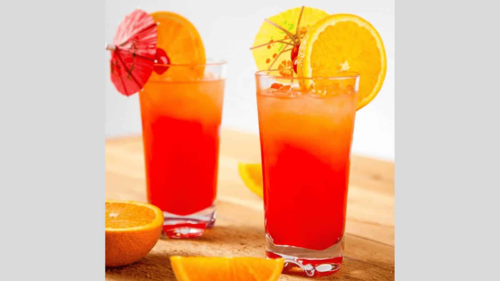 Two tall glasses filled with an orange-red beverage sit on a wooden surface. Each glass is garnished with an orange slice and a cocktail umbrella. Additional orange slices are placed around the glasses.
